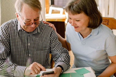 Smiling mature female social worker looking at retired senior man using smart phone in nursing home