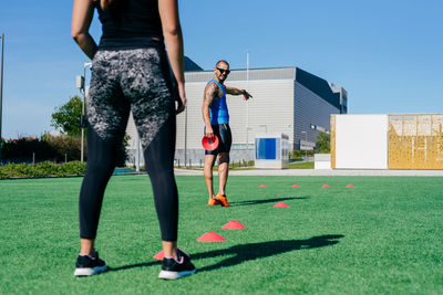 Crop young athletic female performing agility exercise with training cones during fitness workout with personal trainer on sports ground