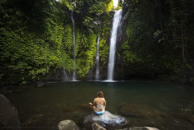 Rear view of woman sitting on rock against waterfall in forest