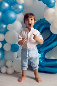 Baby boy in blue pants and shirt standing on the floor in blue and white balloons