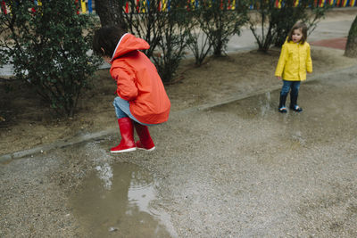 Adorable joyful children in red and yellow raincoat and rubber boots having fun playing in puddle in street in park in gray day