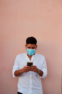 Young latin man with mask is looking his phone on pink background