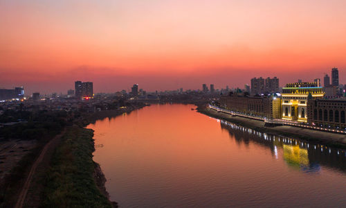 River by illuminated buildings against sky at sunset