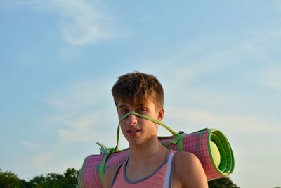 Portrait of young man carrying exercise mat against sky