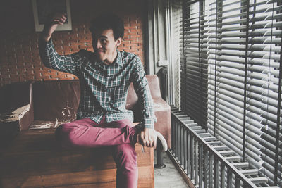 Smiling young man looking away while sitting by window blinds at home