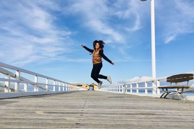 Woman jumping on footbridge against sky