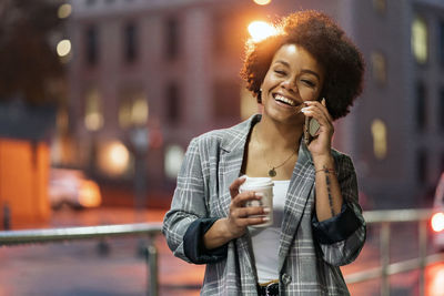 Young woman holding coffee cup talking on phone against building