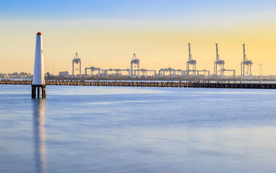 View of lighthouse and container cranes by sea against sky during sunset