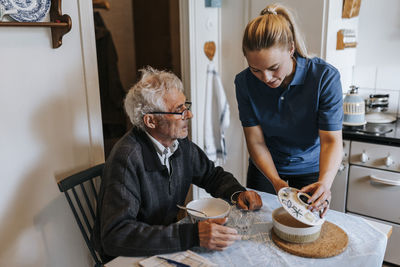 Female caregiver serving food to senior man sitting at dining table in home