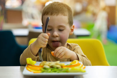 A funny cute three year old toddler boy eats fresh fruit while sitting at a table in a public place.