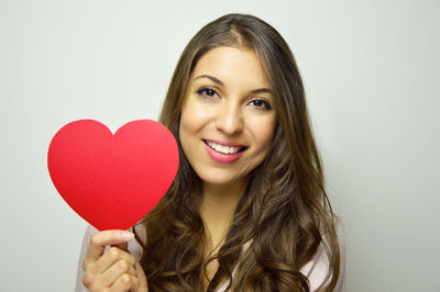Portrait of beautiful young woman holding heart shape paper against gray background