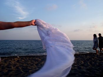 Cropped hand holding white scarf while people walking on shore at beach against sky during sunset