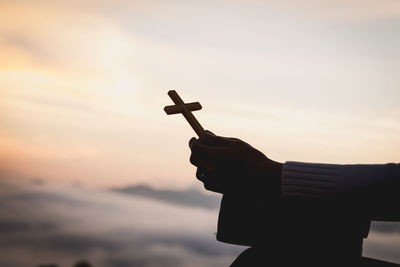 Silhouette hand holding cross against sky during sunset