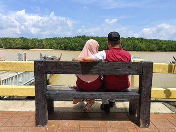 Rear view of couple sitting on bench by lake against sky