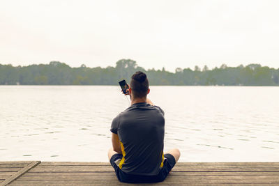 Rear view of man taking selfie while sitting on pier over river against sky