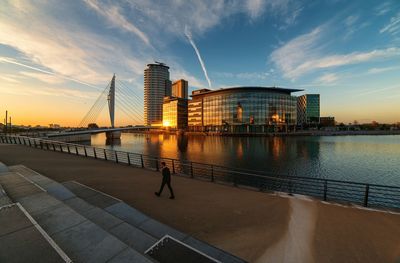 Man walking by river and modern building against sky at sunset