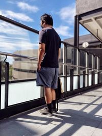 Rear view of young man standing at railings against architecture and sky.