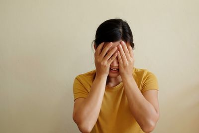 Smiling young woman covering eyes against wall