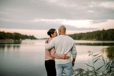 Rear view of smiling couple with arm around standing against lake during sunset