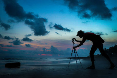 Silhouette man adjusting mobile phone on tripod at shore against cloudy sky