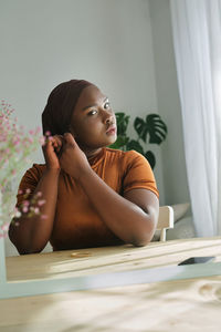 Young african female in traditional headscarf looking in mirror and putting on earrings in modern bedroom