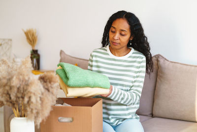 African-american woman packing clothing for charity donation in cardboard box in living room
