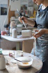 Young craftswoman paints vase with paintbrush after baking and handmade shaping in studio