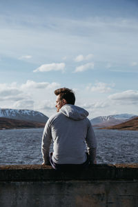 Rear view of man sitting on retaining wall by lake against sky