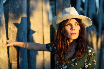 Thoughtful woman wearing hat while standing against wall during sunset