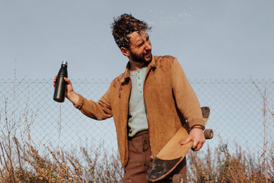 Young bearded male skater with closed eyes in casual outfit pouring water from bottle on head standing near fence in countryside