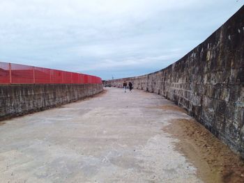 Rear view of people walking on footpath by wall against sky