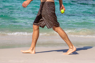 Low section of wet man holding ball while walking on shore at beach during sunny day