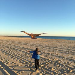 Rear view of boy flying kite at beach against clear sky