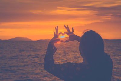 Woman making heart shape towards sun while standing by sea during sunset