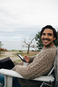 Smiling man with laptop and mobile phone looking away while sitting on chair against sky