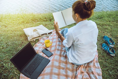 Rear view of woman reading book while by laptop at lakeshore