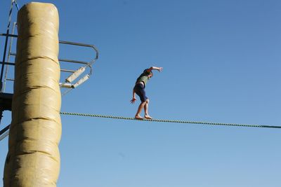 Low angle view of boy walking on rope against blue sky