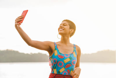 Cheerful millennial female in colorful summer outfit taking selfie on smartphone while enjoying vacation on beach in costa rica