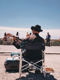 Rear view of man playing guitar while sitting on chair against blue sky during sunny day