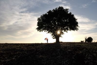 Mid distance view of person holding umbrella by tree on field against sky during sunset