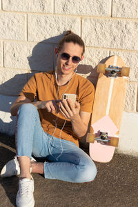 Smiling young man with surf skate on the side sitting on the floor chatting on smartphone having fun