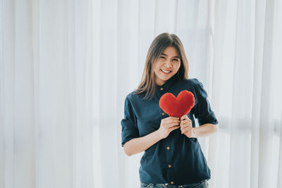 Portrait of smiling woman holding heart shape while standing against window