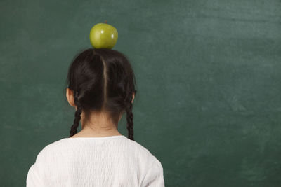 Rear view of girl with granny smith apple on head standing against wall