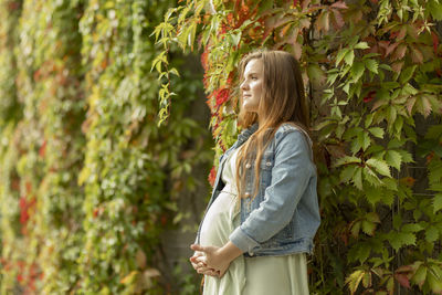 White pretty pregnant woman stand near green overgrown liana wall. long brown haired young healthy
