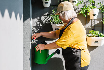 Side view of elderly female gardener in apron and cap refilling watering can in a balcony at home on a sunny day