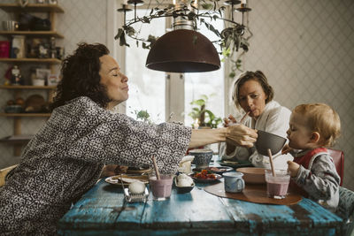 Mature woman feeding water to daughter sitting by girlfriend at dining table