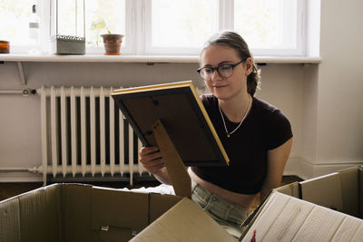 Woman holding picture frame amidst cardboard boxes at home