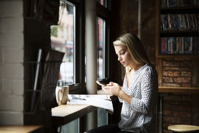 Young woman using phone while reading newspaper at table in cafe