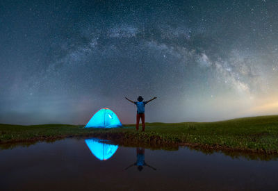 Rear view of mature man with arms outstretched standing on grassy field by lake at campsite against star field