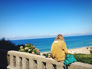 Rear view of woman sitting on stone railing against blue sky